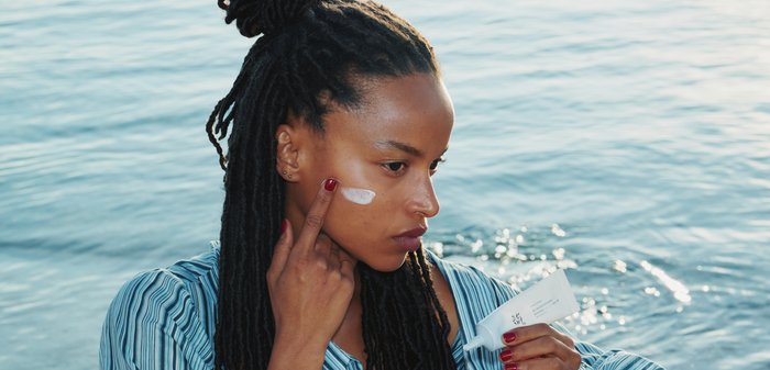 Femme aux cheveux tressés appliquant une crème blanche sur sa joue près d'un plan d'eau, tenant un tube de produit de soin de la peau.