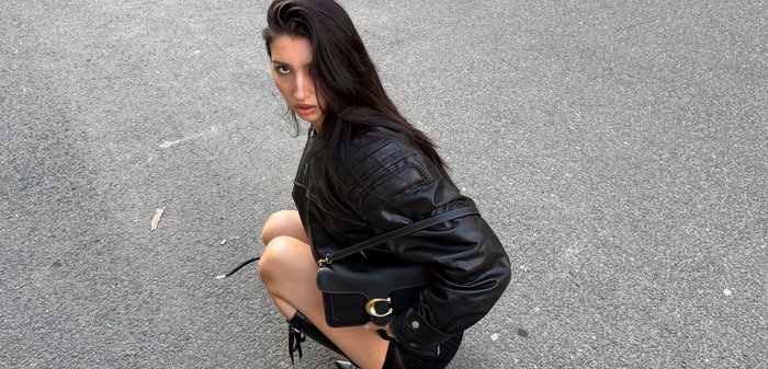 Junge Frau mit langen dunklen Haaren, die auf Asphalt hockt, trägt eine schwarze Lederjacke, einen schwarzen Minirock, spitze schwarze Stiefel und hält eine schwarze Handtasche.