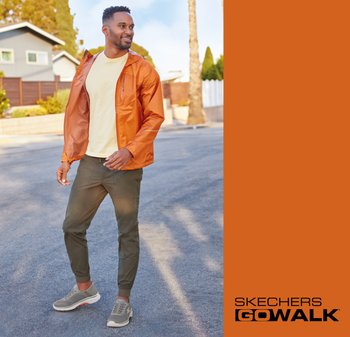 Skechers GOwalk footwear displayed with an orange windbreaker jacket, yellow shirt, and olive green joggers on a paved street.