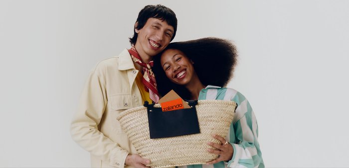 Deux personnes souriantes tiennent un grand panier tressé contenant une enveloppe orange Zalando sur un fond blanc uni.