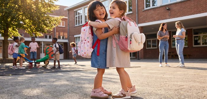 Due giovani ragazze con zaini sorridono e si abbracciano in un cortile della scuola, con altri bambini e un insegnante sullo sfondo.