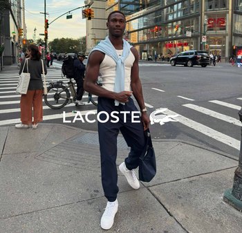 Camisa blanca sin mangas, suéter azul claro atado alrededor de los hombros, pantalones azul marino y zapatillas blancas; bolso negro y fondo urbano.