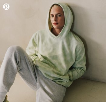 Giovane seduto contro un muro che indossa una felpa con cappuccio verde chiaro tie-dye e pantaloni della tuta grigi, che guarda direttamente l'obiettivo.