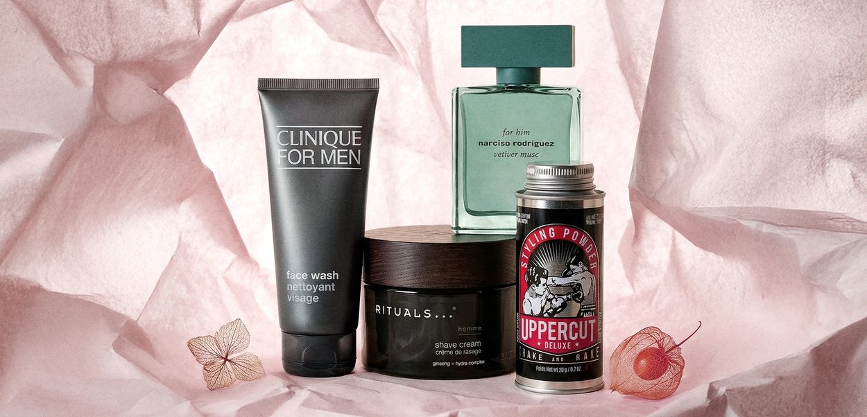 Clinique żel do mycia twarzy, krem do golenia Rituals, woda kolońska Narciso Rodriguez oraz pudrowa pasta do stylizacji Uppercut Deluxe na różowym tle o fakturze.