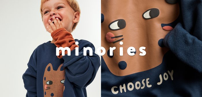 Nuori lapsi, jolla on lyhyet vaaleat hiukset, nauraa käden osittain peittäessä suuta, pukeutunut tummansiniseen neuleeseen, jossa on ruskea kissakuva ja oranssit hihat.