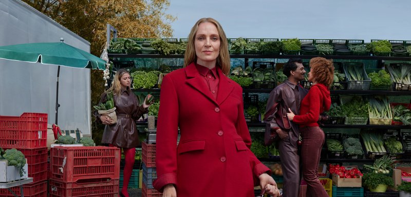 Manteau rouge avec des boutons, porté sur une chemise rouge. Veste en cuir marron et parapluie vert à proximité. Paniers empilés avec des légumes frais en arrière-plan.