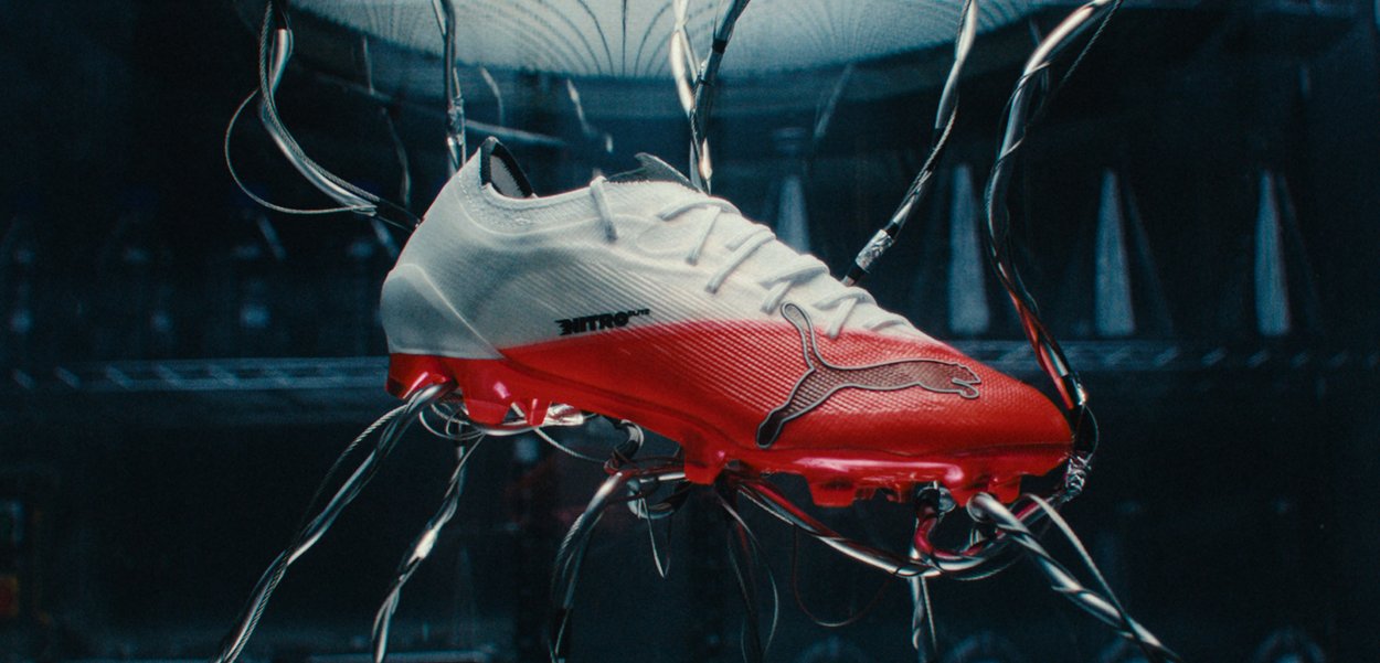 Bota de fútbol Puma blanca y roja sostenida por varios brazos robóticos en un laboratorio oscuro y de alta tecnología.