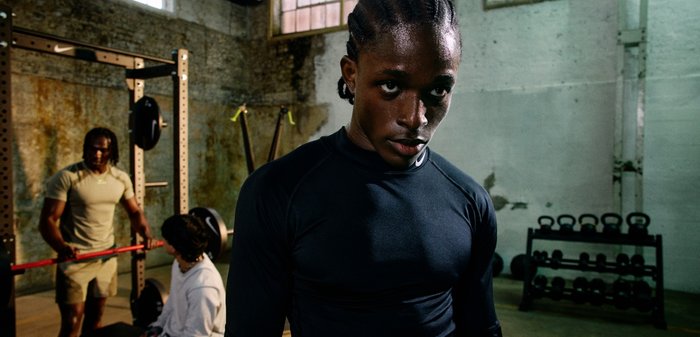 Black athletic long-sleeve top with a fitted design. The background features gym equipment, including a squat rack and weights.