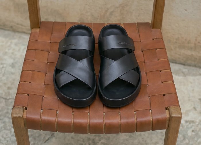 Een paar zwarte lederen sandalen met gekruiste bandjes geplaatst op een bruine gevlochten leren stoel tegen een neutrale achtergrond.