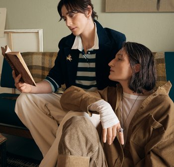 Due persone sedute su un divano verde. Una legge un libro, indossando un blazer blu scuro e una camicia a righe. L'altra con una giacca di velluto a coste beige e una maglietta bianca.