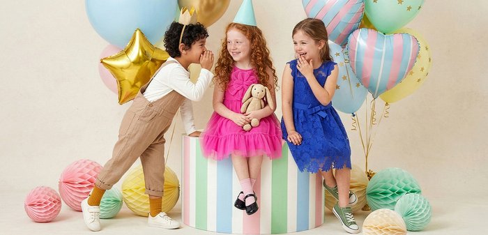 Trois enfants à une fête d'anniversaire, l'un chuchotant à une fille en rose tenant un lapin en peluche, avec des ballons colorés et des décorations autour d'eux.