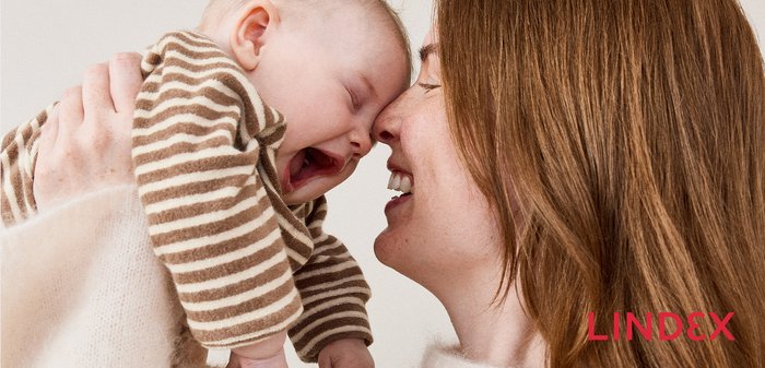 Femme souriante touchant le nez d’un bébé riant portant une chemise rayée marron et blanche, tous deux vêtus de vêtements doux et clairs.