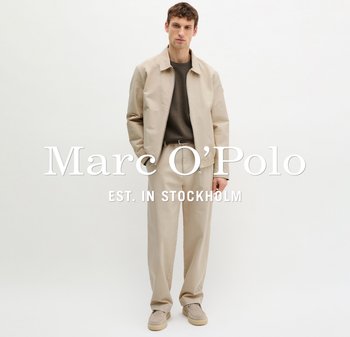 Młody mężczyzna w beżowej kurtce i spodniach, ciemnobrązowej koszuli i beżowych butach, stojący na tle białej, jednolitej ściany.