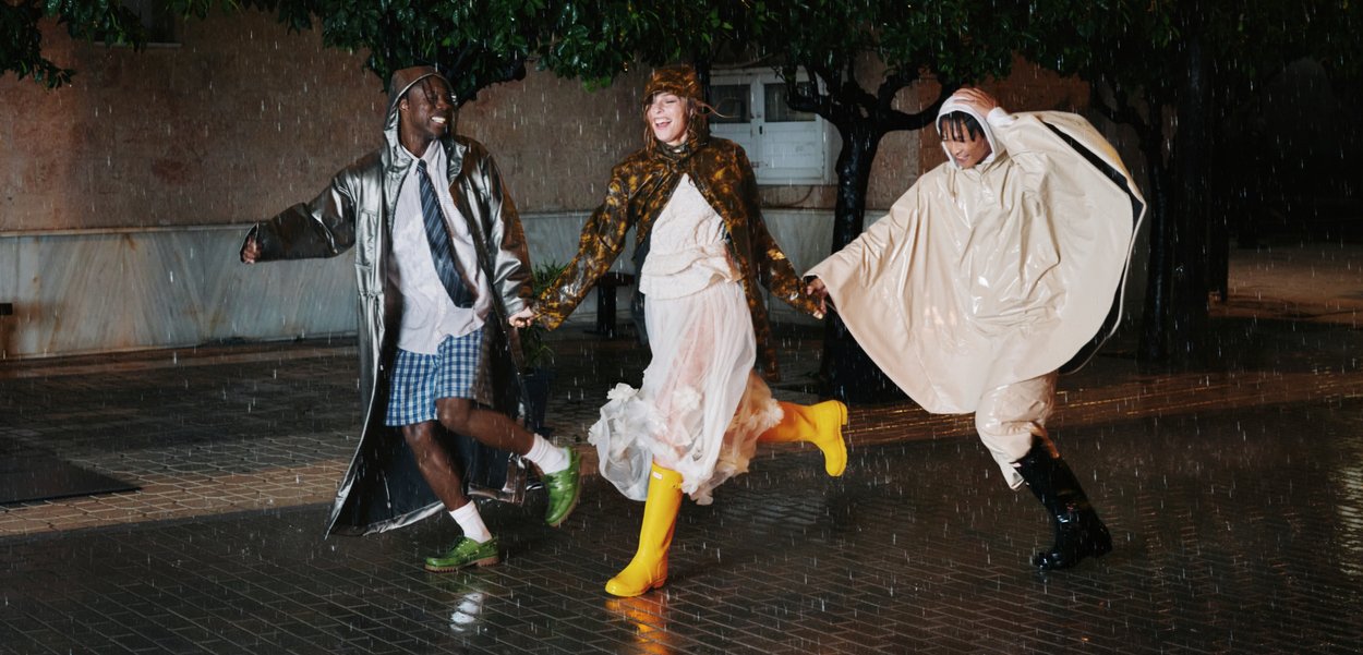 Three people wearing raincoats and wellies dance joyfully on a wet street at night under heavy rain.