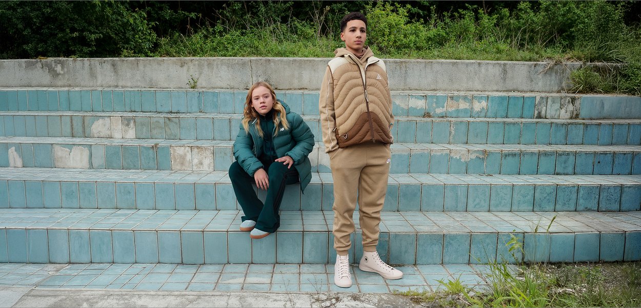 Två personer sitter på en trappa med blå kakelplattor. Den ena bär en tealfärgad isolerad jacka och matchande byxor; den andra bär en beige vadderad jacka och beige byxor.