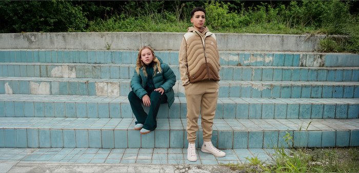 Deux personnes sont assises sur un escalier aux carreaux bleus. L'une porte une veste isolante bleu sarcelle et un pantalon assorti ; l'autre porte une veste matelassée beige et un pantalon beige.