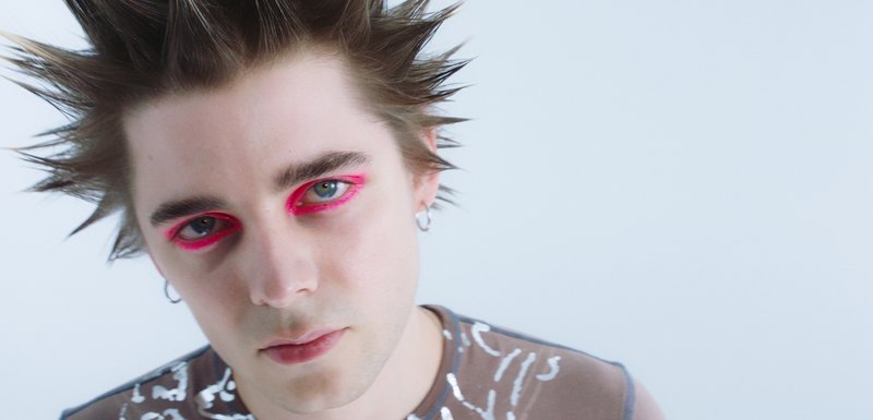 Portrait d'une personne avec des cheveux en piques, portant une chemise brune avec des motifs blancs et un maquillage des yeux rose vif. Fond simple, tons neutres.