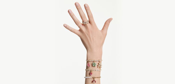 Hand with three decorative rings and three charm bracelets featuring watermelon, clover, bear, strawberry, and bee charms.