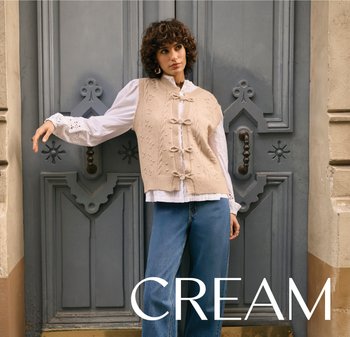 Beige Strickweste mit strukturiertem Muster und Schleifendetails über einem weißen Hemd, kombiniert mit hellblauen Jeans mit weitem Bein, vor einer grauen Tür.