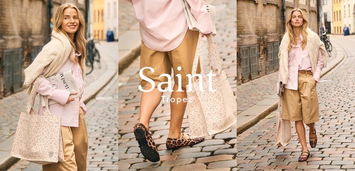 Blonde woman wearing a pink shirt, beige shorts, and a cream sweater walks on a cobblestone street in an urban setting.