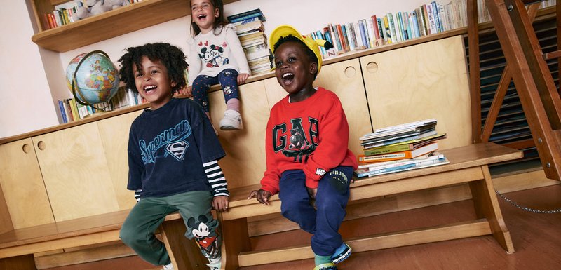 Trois enfants sont assis sur un banc en bois. Le garçon à gauche porte un t-shirt Superman bleu marine et un pantalon vert ; la fille au centre a un haut Disney et des leggings à motifs. Le garçon à droite porte un hoodie rouge GA et un pantalon de jogging bleu marine.