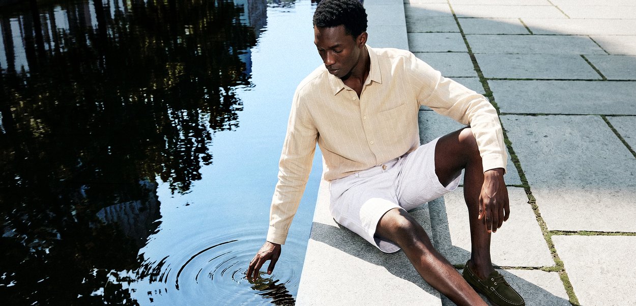 Hombre con camisa beige y pantalones cortos blancos sentado en un borde de piedra, tocando suavemente el agua en un canal o estanque tranquilo al aire libre.