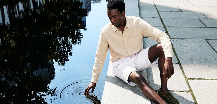 Hombre con camisa beige y pantalones cortos blancos sentado en un borde de piedra, tocando suavemente el agua en un canal o estanque tranquilo al aire libre.
