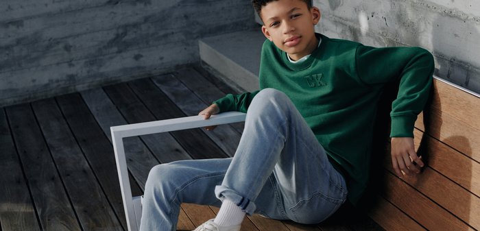 Adolescent garçon en sweat-shirt vert et jean bleu clair assis sur un sol en bois contre un mur en béton, portant des baskets blanches Calvin Klein.