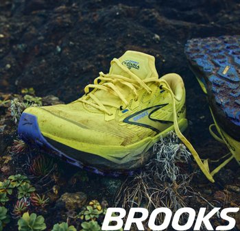 Brooks Cascadia trailrunschoen, gele mesh bovenkant, blauwe accenten, gestructureerde buitenzool, zichtbare vuiligheid, veters en verstevigde neuskap.