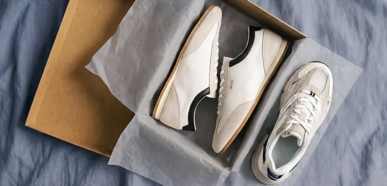 Par de zapatillas blancas y beige en una caja de zapatos marrón abierta, con una zapatilla colocada fuera sobre una superficie de tela azul.