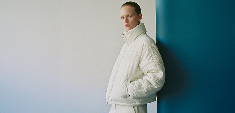 Veste blanche bien rembourrée avec un col haut, présentant des sections cousues et une fermeture éclair, sur un fond bleu et blanc.