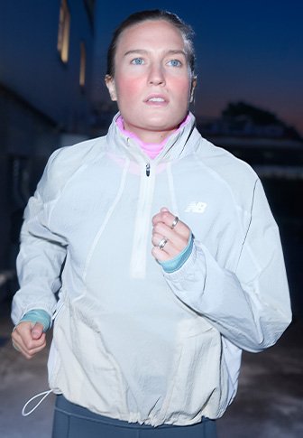 Ženska teče na prostem ob mraku, oblečena v belo jakno New Balance in osredotočena na svoj tek.