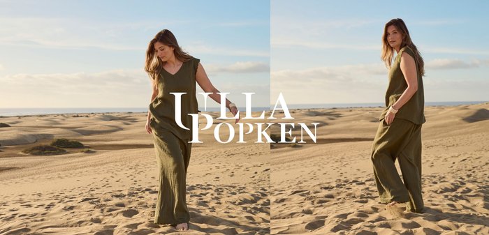 Woman in loose olive-green outfit walking barefoot on sandy dunes under a clear sky, with distant ocean horizon.