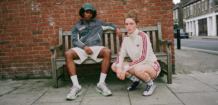 Deux jeunes en tenue sportive, l'un assis sur un banc en bois, l'autre accroupi sur un trottoir près d'un mur en briques.