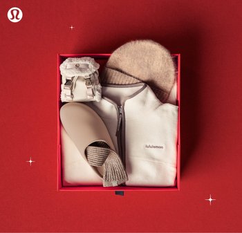 Rode cadeaubox met een beige fleecejack, bijpassende beanie, geribbelde sokken in de slipper en een kleine harige rugzak op een rode achtergrond.