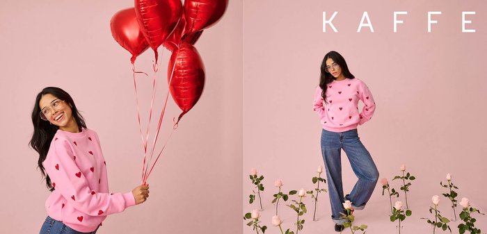 Woman in pink heart-patterned sweater and jeans holds red heart-shaped balloons and poses among pink roses against a soft pink background.