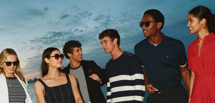 Sechs junge Erwachsene in stylischer Freizeitkleidung und Sonnenbrillen stehen im Freien bei Sonnenuntergang in der Nähe von Wasser und lächeln.