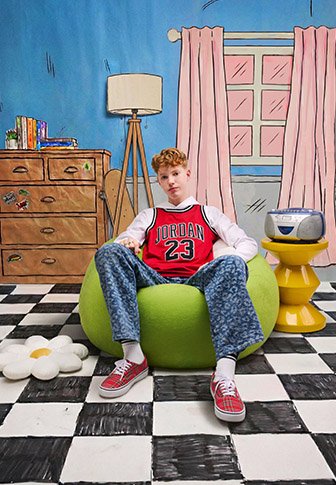 Adolescent portant un maillot rouge Jordan et un pantalon à motifs, assis sur un pouf vert dans une pièce colorée avec un sol à carreaux et une décoration rétro.