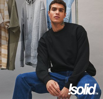 Schwarzer Sweatshirt mit Rundhalsausschnitt aus Baumwollmaterial. Modell sitzt mit blauen Jeans. Im Hintergrund hängen gestreifte und einfarbige Hemden.