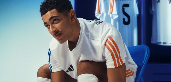 Joueur de football masculin en maillot blanc avec des rayures orange et bleu attachant ses crampons dans le vestiaire avec des maillots numérotés accrochés.