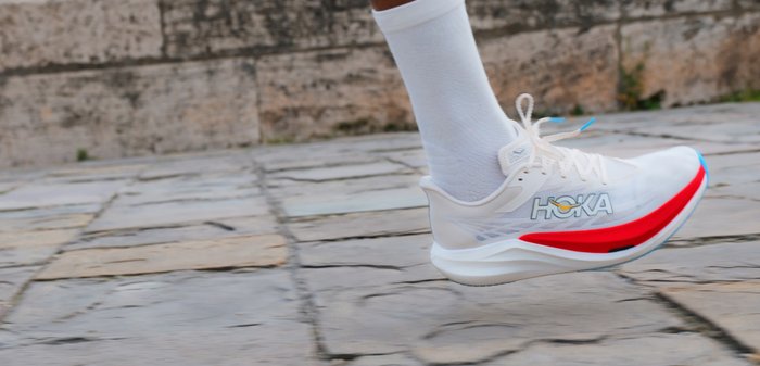 Πόδι δρομέα στον αέρα, φορώντας λευκό αθλητικό παπούτσι Hoka με κόκκινες και μπλε λεπτομέρειες, σε υπόβαθρο λιθόστρωτου δρόμου.