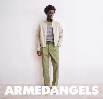 Uomo con giacca beige, camicia a righe verde, pantaloni oliva, in piedi contro un muro bianco con testo "ARMEDANGELS" sotto.