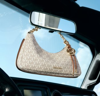 Bolso beige con monograma de Michael Kors con asa de cadena dorada colgando del espejo retrovisor de un coche contra un fondo de cielo azul.