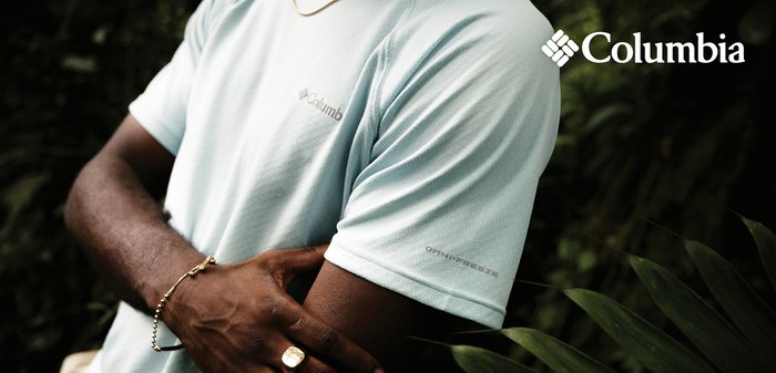 Dark-skinned person wearing light blue Columbia Omnifreeze shirt, gold ring, and bracelet, standing near green leaves outdoors.