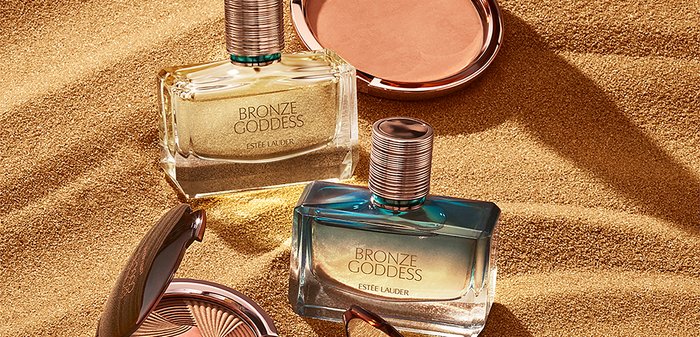 Deux flacons Bronze Goddess d'Estée Lauder : l'un rectangulaire avec un design doré et transparent, l'autre en dégradé de bleu, à côté d'une poudre bronzante.