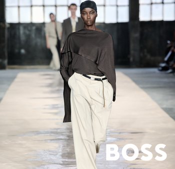 Brun draperet top, beige højtaljede bukser med bælte. Model der går på en catwalk med en tekstureret grå overflade. Industrielt baggrund.