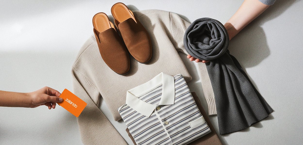 Braune Slipper, beiger strukturierter Pullover, gestreiftes Polohemd, grauer Schal und eine orangefarbene Zalando-Geschenkkarte auf einem neutralen Hintergrund arrangiert.