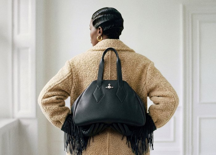 Άτομο που φοράει μπεζ παλτό με υφή, κρατώντας μαύρη δερμάτινη τσάντα με ασημένιο λογότυπο στο πίσω μέρος, στέκεται σε φωτεινό εσωτερικό χώρο.
