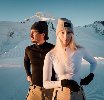 Zwei Personen stehen in einer verschneiten Landschaft, tragen enganliegende Thermoshirts, hellbeige Hosen und Mützen. Ein Shirt ist schwarz, das andere weiß.