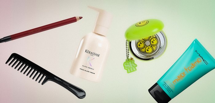 Crayon contour des lèvres, flacon de crème capillaire, peigne à dents larges, compact vert avec visage souriant et boutons, et tube bleu de mousse coiffante sur une surface pâle.